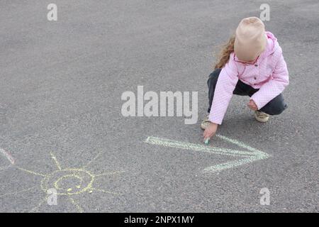 Una bambina in una giacca e cappello sulla strada disegna una freccia con gesso sul marciapiede. Bambini che giocano all'aperto in una giornata di sole. Foto Stock