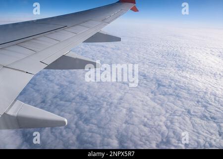 Volando sull'aeroplano sopra le nuvole puffy sotto l'ala attraverso la finestra dell'aereo di un aeroplano. Vista aerea dalla finestra dell'aereo. Foto Stock