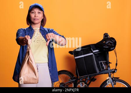 Serio dipendente asiatico consegna ristorante che fa segno di disapprovazione mentre consegna sacchetti di cibo da portare via ai clienti. Operaio pizzeria che porta gli ordini con la bicicletta. Servizio di ristorazione e concetto Foto Stock