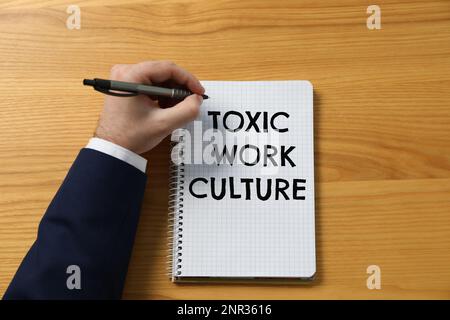 Uomo scrivere testo Toxic Work Cultura in notebook a tavola di legno, vista dall'alto Foto Stock