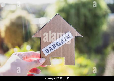 Etichetta di affare sulla casa di cartone di fronte al bokeh del cortile, concetto di trovare una proprietà di sogno e di mercato immobiliare abbordabilità Foto Stock