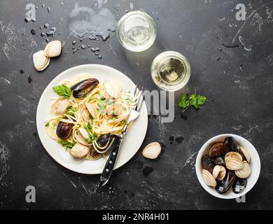 Spaghetti alle vongole, pasta italiana di pesce con vongole e cozze, in piatto con erbe e due bicchieri di vino bianco su fondo rustico in pietra. Foto Stock