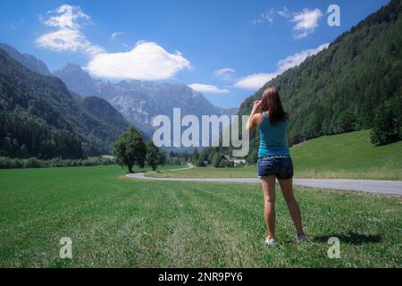 Donna sconosciuta vista da dietro fotografando la magica valle di Logarska. Bellissimo turista a Logarska dolina scattare una foto. Foto Stock