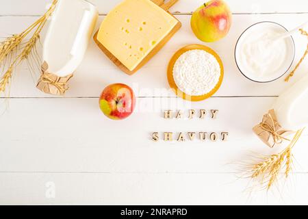 diversi tipi di latticini in ciotole, bottiglie su fondo bianco. vista dall'alto. giacitura piatta. lettere di legno con il testo shavuot Foto Stock