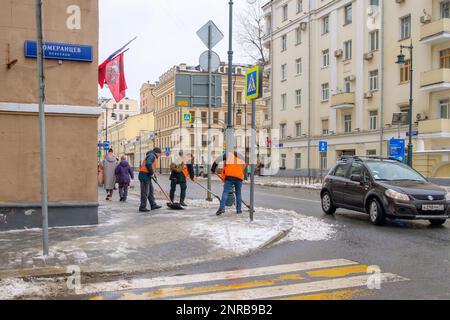 Mosca. Russia. Febbraio 25, 2023. I lavoratori in giubbotti arancioni spazzano la neve al largo del marciapiede su una strada cittadina in una nuvolosa giornata invernale. Foto Stock