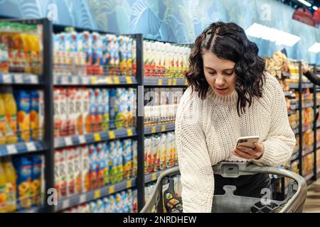 una donna mette un prodotto con un elenco di prodotti sul telefono in un carrello nel supermercato Foto Stock