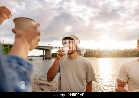 Giovane uomo sorridente che indossa un cappello da secchio e che si diverti a bere con un amico durante il picnic Foto Stock