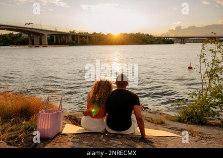 Vista posteriore della coppia guardando il tramonto mentre si siede su coperta da picnic vicino al mare Foto Stock