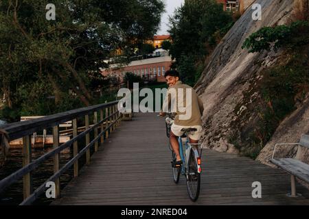 Felice giovane uomo che guarda oltre le spalle mentre pedala sul molo Foto Stock