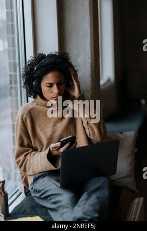 Imprenditore donna che indossa cuffie wireless e lavora su un computer portatile dalla finestra in ufficio creativo Foto Stock