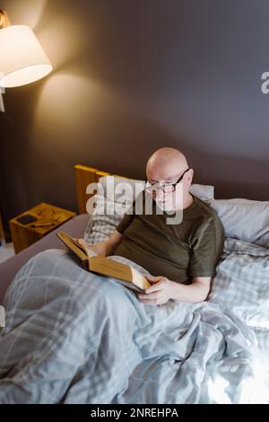Uomo anziano che indossa gli occhiali mentre legge il libro sul letto a casa Foto Stock