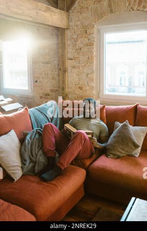 Un giovane uomo che legge un libro mentre si rilassa sul divano a casa Foto Stock