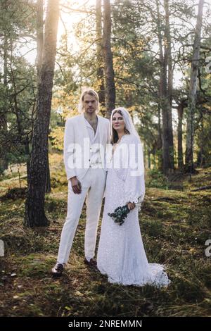 Ritratto di sposa ben vestita e sposo in piedi da alberi nella foresta al matrimonio Foto Stock