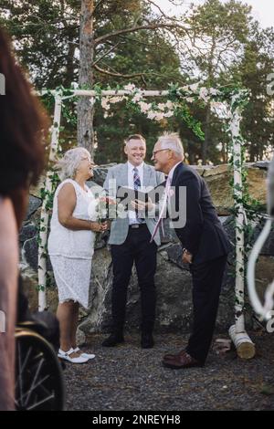 Coppia anziana scambiando voti dal ministro in piedi contro il muro con decorazione floreale durante la cerimonia nuziale Foto Stock