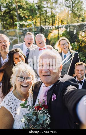 Giovane coppia giovane allegra che prende selfie con la famiglia e gli amici al matrimonio Foto Stock