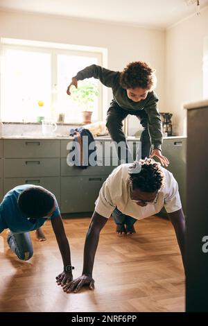 Figlio giocoso in piedi sulla schiena del padre facendo push-up in cucina a casa Foto Stock