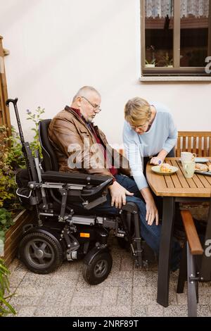 Donna anziana che assiste l'uomo in sedia a rotelle motorizzata con disabilità in cortile Foto Stock