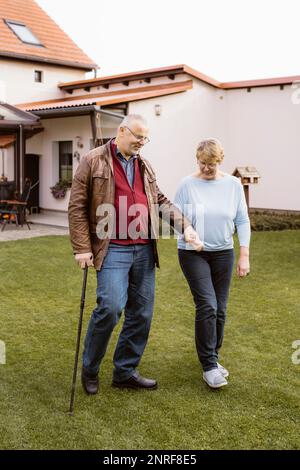 Uomo anziano che tiene la mano della donna mentre cammina con la canna al prato nel cortile posteriore Foto Stock