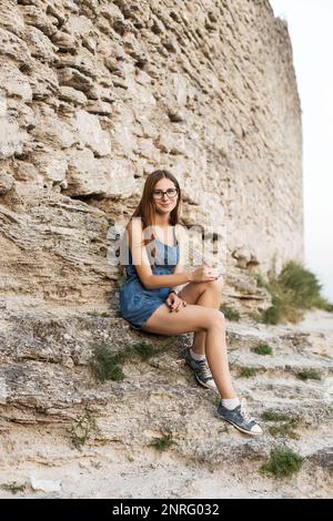 donna in denim tuta si siede vicino a muro di pietra sulla montagna vicino alla città Foto Stock