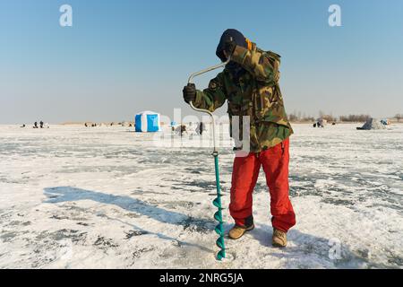 Un ragazzo asiatico vestito calorosamente trapana un buco nel ghiaccio sul Fi inverno Foto Stock