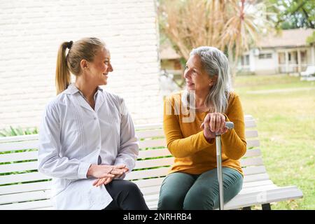 Donna anziana che parla con l'infermiera femminile sul banco in giardino di casa di cura Foto Stock