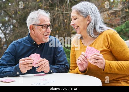 Felice uomo anziano e donna godendo mentre giocando le carte a tavolo nel giardino di casa di cura Foto Stock