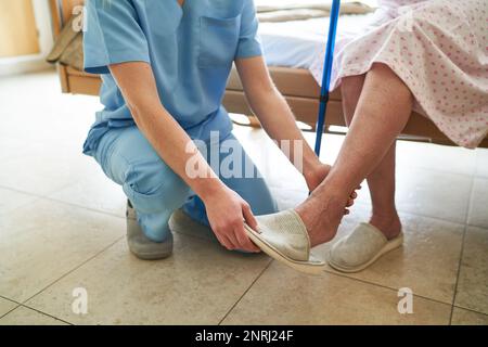 Bassa sezione dell'infermiera che aiuta la donna anziana nel mettere sulle pantofole nella casa di cura Foto Stock