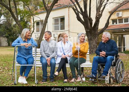Medico felice che parla con un gruppo di persone anziane sedute su panchina in giardino di casa di cura Foto Stock
