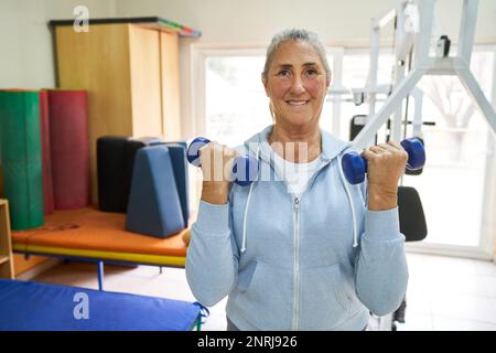 Ritratto di donna anziana sorridente che si esercita con manubri mentre si trova in piedi in classe di fitness Foto Stock