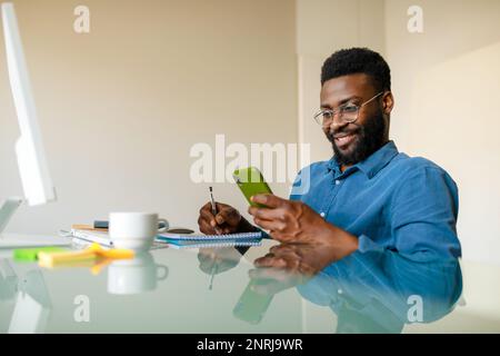 Felice imprenditore nero maschio seduto alla scrivania, guardando il cellulare, guardando il webinar e prendendo appunti, spazio libero Foto Stock