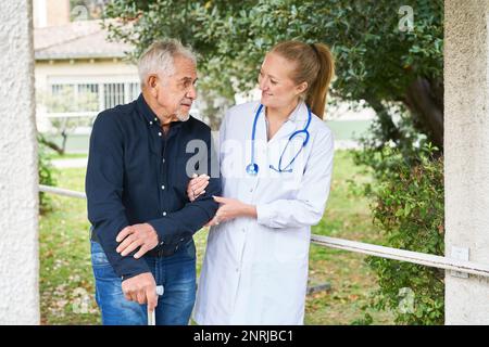 Sorridente giovane dottoressa che sostiene l'uomo anziano disabile a camminare in giardino Foto Stock