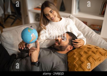 Felice uomo arabo e giovane uomo pensivo europeo riposo sul divano, tenere il globo, scegliere il paese per la vacanza Foto Stock