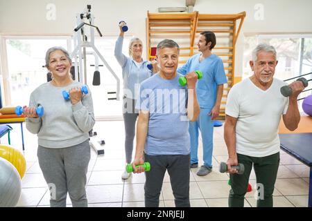 Gli uomini anziani e la donna che esercitano con i dumbbells mentre il terapeuta che assiste il paziente in background nella casa di cura Foto Stock