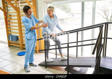 Un caregiver maschile assiste una donna anziana sorridente sulla rampa durante la terapia del movimento presso il centro di riabilitazione Foto Stock