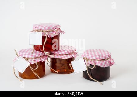 Una serie di vasetti per marmellata con coperchi a scacchi rosa e bianchi ed etichette vuote isolate su sfondo bianco Foto Stock