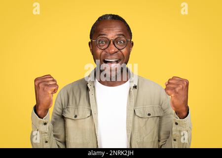 Felice felice eccitato ragazzo nero maturo in casual e occhiali alza le mani, celebrando la vittoria Foto Stock