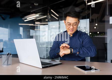 Un uomo d'affari asiatico di successo usa l'orologio intelligente sul posto di lavoro, l'uomo sceglie le app e sorride felice con le app, i dipendenti in ufficio usano il notebook al lavoro. Foto Stock