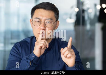 Primo piano ritratto di un uomo che tiene il dito vicino alla bocca chiedendo silenzio sul posto di lavoro, l'uomo asiatico in ufficio guardando la fotocamera turbato e preoccupato cercando di concentrarsi. Foto Stock