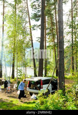 Un uomo cammina tra un'auto e una tenda turistica in un parcheggio nella foresta nelle montagne Altai vicino al fiume. Foto Stock