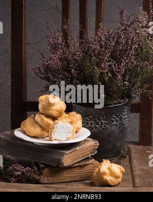 Autunno Vintage Provence Still Life con un gruppo di dolci francesi Crema bianca Eclairs sul piatto, fiori di Heather e vecchi libri. Cemento grigio rustico Ru Foto Stock