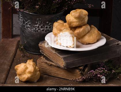 Autunno Vintage Provence Still Life con un gruppo di dolci francesi Crema bianca Eclairs sul piatto, fiori di Heather e vecchi libri. Cemento grigio rustico Ru Foto Stock