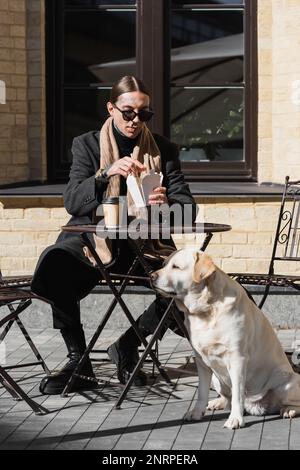 lunghezza completa di tatuato uomo in occhiali da sole tenendo bastoncini mentre mangiare cibo asiatico vicino al cane in caffè all'aperto, immagine stock Foto Stock