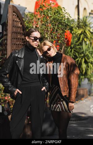 donna bionda in occhiali da sole appoggiata sulla spalla di un uomo elegante posa con le mani in tasche, immagine stock Foto Stock