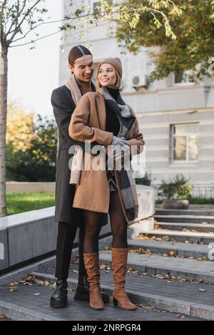 tutta la lunghezza dell'uomo tatuato felice che abbraccia la donna bionda nel cappello mentre si levano in piedi fuori, immagine di scorta Foto Stock