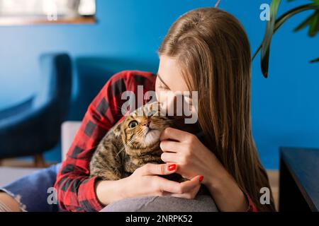 Bella giovane donna sorridente che tiene Scottish Tabby Cat siede in Couch a casa, il concetto di amare e prendersi cura degli animali domestici Foto Stock