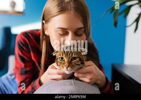 Bella giovane donna sorridente che tiene Scottish Tabby Cat siede in Couch a casa, il concetto di amare e prendersi cura degli animali domestici Foto Stock