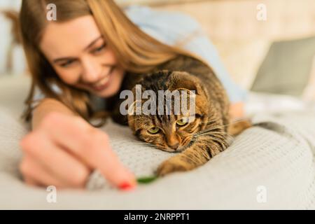 Sorridente giovane donna bionda sdraiata nel suo letto in camicia una carina domestica giocando gatto scozzese tabby a casa, concetto di amare e prendersi cura degli animali domestici Foto Stock