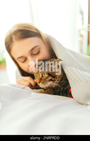 Cute sorridente giovane donna bionda sdraiata su un letto giocando un carino Green-eyed Scottish Tabby Cat, il concetto di amare e prendersi cura degli animali domestici, adottandoli Foto Stock