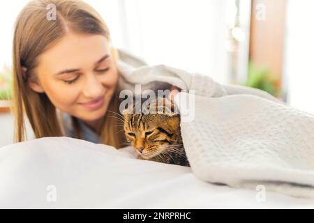 Cute sorridente giovane donna bionda sdraiata su un letto giocando un carino Green-eyed Scottish Tabby Cat, il concetto di amare e prendersi cura degli animali domestici, adottandoli Foto Stock