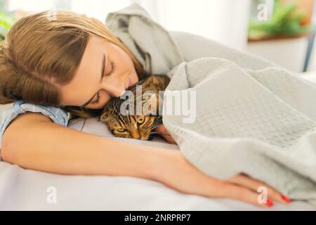 Cute sorridente giovane donna bionda sdraiata su un letto giocando un carino Green-eyed Scottish Tabby Cat, il concetto di amare e prendersi cura degli animali domestici, adottandoli Foto Stock